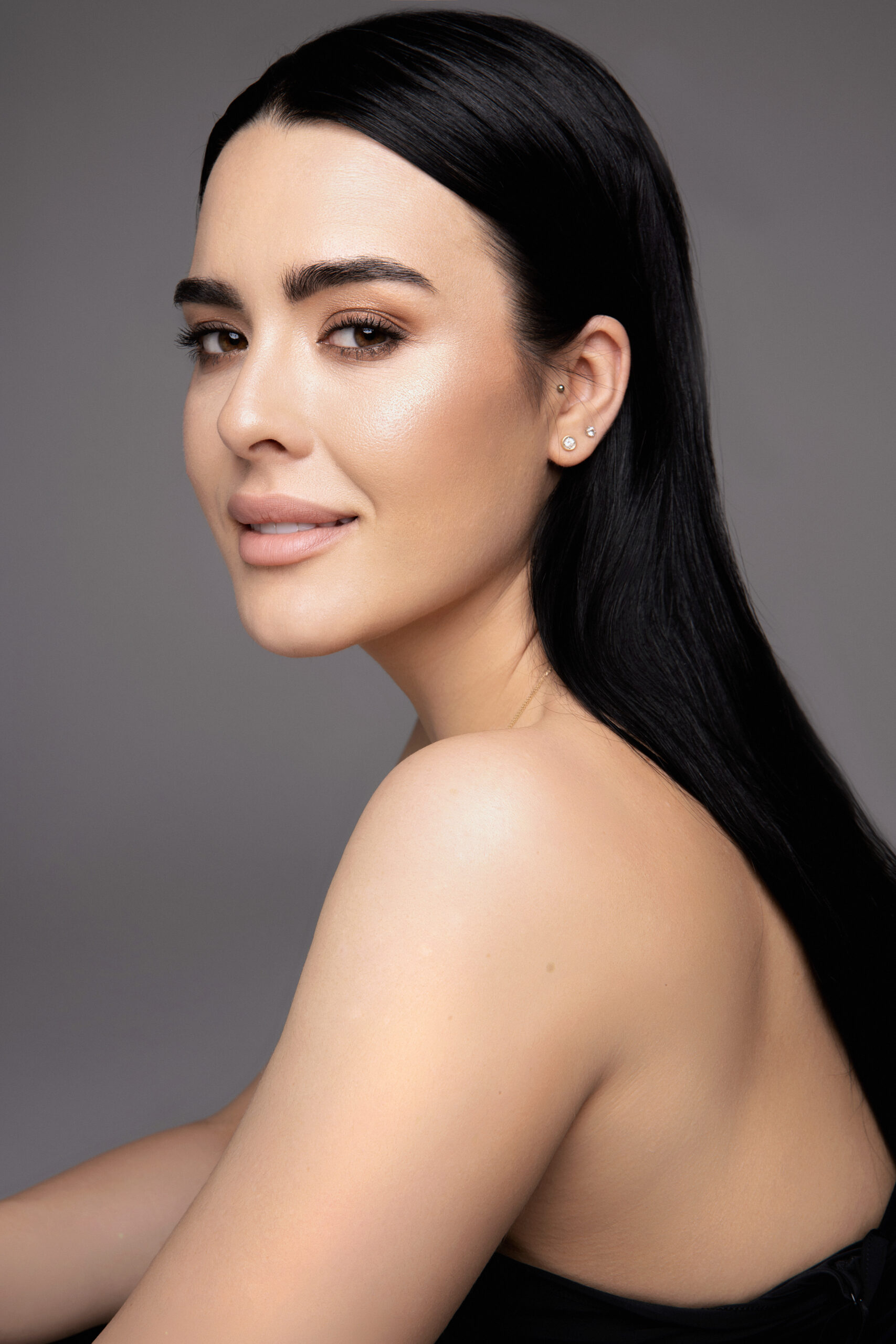 Kobieta o długich, prostych czarnych włosach i promiennej cerze pozuje w staniku bez ramiączek na szarym tle, a jej metamorfoza uśmiechu jest podkreślona, gdy uśmiecha się delikatnie i patrzy przez ramię do kamery.