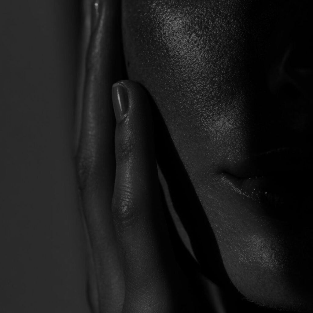 Zbliżenie czarno-białego zdjęcia twarzy osoby częściowo w cieniu, z jedną ręką delikatnie dotykającą jej policzka. Obraz podkreśla fakturę skóry, tworząc dramatyczną, nastrojową atmosferę przypominającą metamorfozę uśmiechu.